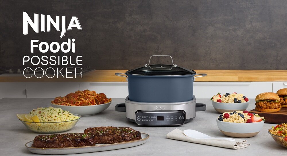 Na blacie kuchennym stoi wolnowar z logo Ninja, otoczony różnorodnymi potrawami. Obok urządzenia widoczny jest napis: \'NINJA Foodi POSSIBLE COOKER\'.