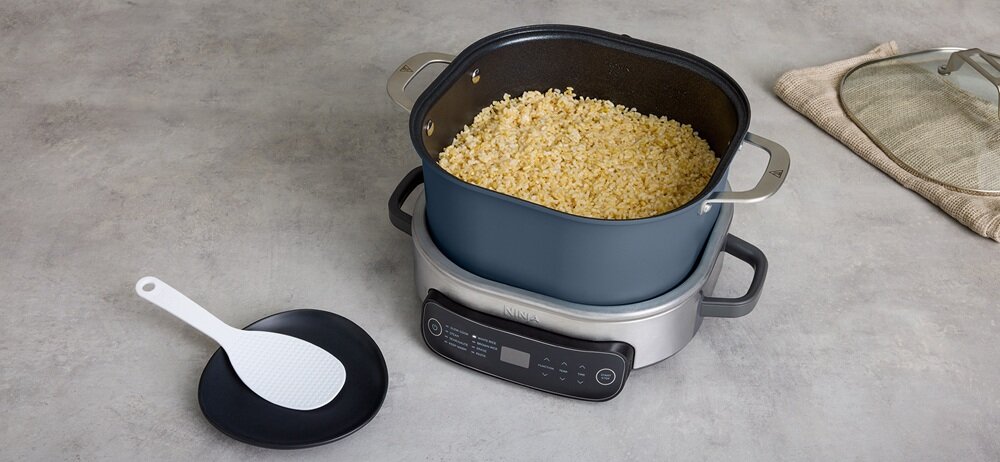 Na blacie stoi wolnowar z gotującym się ryżem, obok leży biała łopatka na czarnym talerzu. W tle widoczna jest pokrywka z uchwytem.