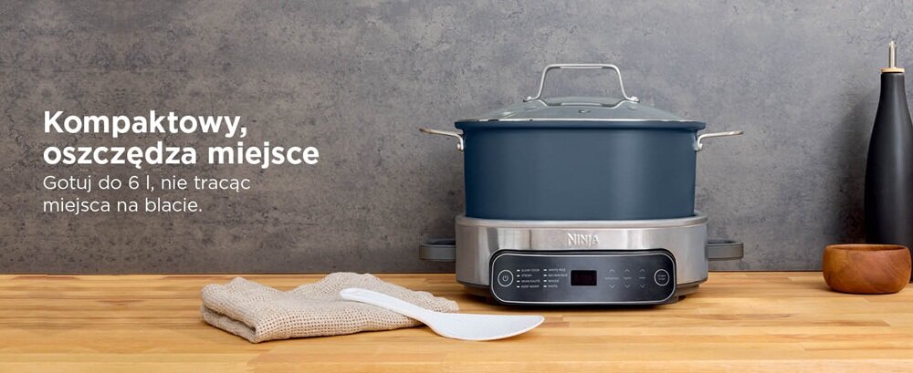 Na drewnianym blacie stoi wolnowar z niebieskim garnkiem i srebrną podstawą z wyświetlaczem. Obok znajduje się ściereczka, łopatka i drewniana miska, a na ścianie widoczny jest tekst: \'Kompaktowy, oszczędza miejsce. Gotuj do 6 l, nie tracąc miejsca na blacie.\'