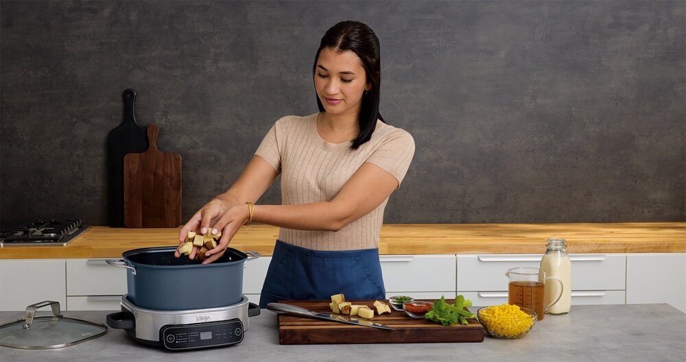 Kobieta wkłada pokrojone ziemniaki do wolnowaru marki Ninja, stojącego na blacie kuchennym. Obok znajdują się deska z warzywami, dzbanek z mlekiem i inne składniki.
