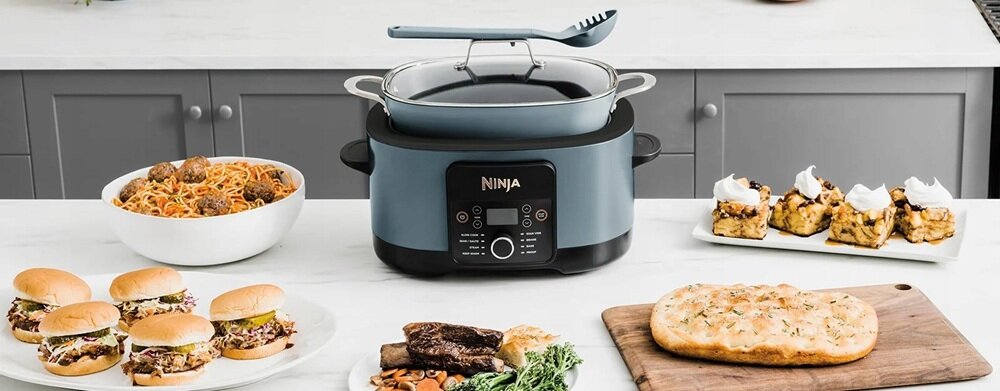 Na blacie kuchennym znajduje się wolnowar z logo \'NINJA\', otoczony różnorodnymi potrawami, w tym burgerami, makaronem i ciastem. W tle widoczne są szafki kuchenne.
