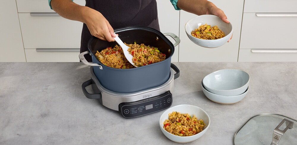 Osoba miesza potrawę w dużym garnku umieszczonym na urządzeniu kuchennym z wyświetlaczem i przyciskami. Obok znajdują się miski z podobną potrawą oraz szklana pokrywka.