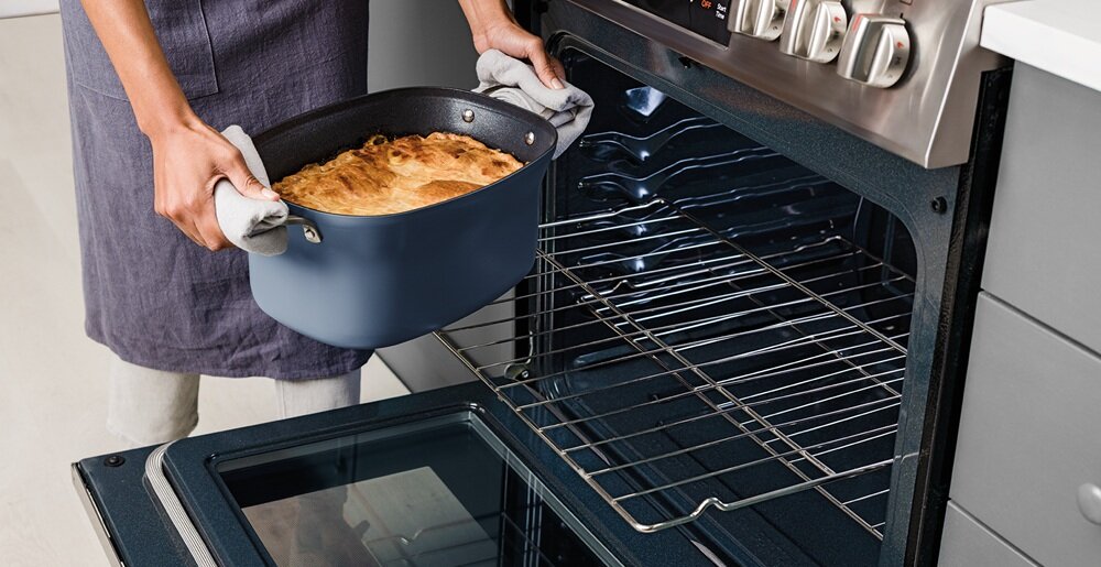 Osoba wyciąga niebieski garnek z piekarnika, trzymając go za pomocą rękawic kuchennych. Wnętrze piekarnika jest puste, a na górnej półce znajduje się potrawa w garnku.