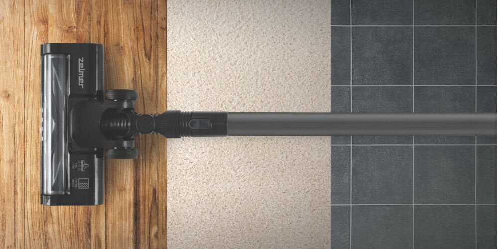 Odkurzacz z logo \'Zelmer\' na głowicy, widoczny na tle trzech różnych powierzchni: drewnianej, dywanowej i kafelkowej.