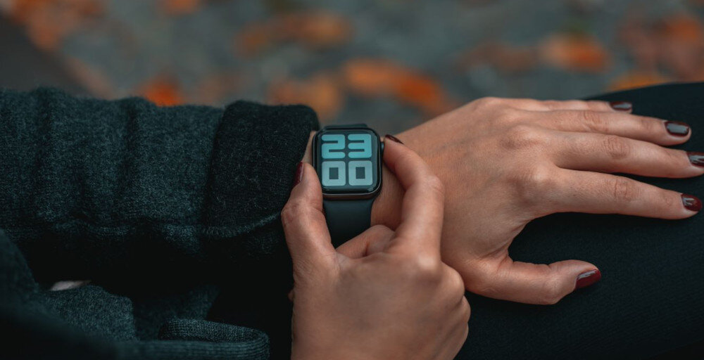 Osoba nosi zegarek z dużym wyświetlaczem cyfrowym pokazującym godzinę 23:00. Tło jest rozmyte, z widocznymi jesiennymi liśćmi.