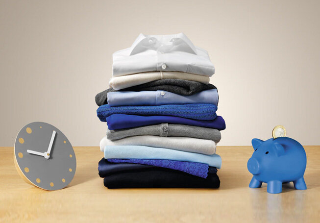 Na drewnianym blacie leży stos złożonych ubrań w różnych odcieniach niebieskiego, szarego i białego. Obok znajduje się niebieska skarbonka w kształcie świnki oraz szary zegar z żółtymi kropkami zamiast cyfr.