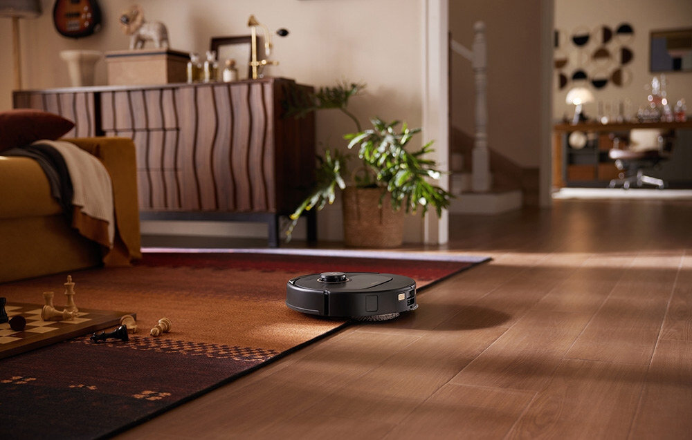Czarny robot sprzątający porusza się po drewnianej podłodze w przytulnym wnętrzu z dywanem i rośliną w tle. W pomieszczeniu widoczne są meble i dekoracje.