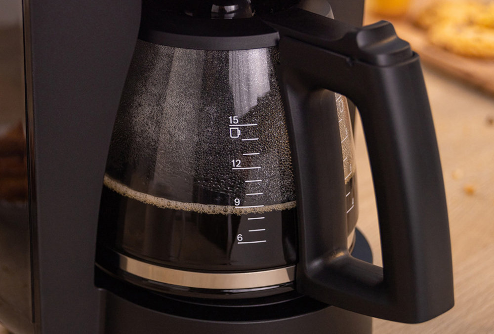 Czarny ekspres do kawy z przezroczystym dzbankiem, na którym widoczne są oznaczenia poziomu wody od 6 do 15. W tle widać rozmyty fragment blatu kuchennego.