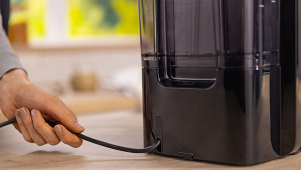 Osoba trzyma kabel podłączony do czarnego urządzenia kuchennego. W tle widoczne są rozmyte elementy kuchni.