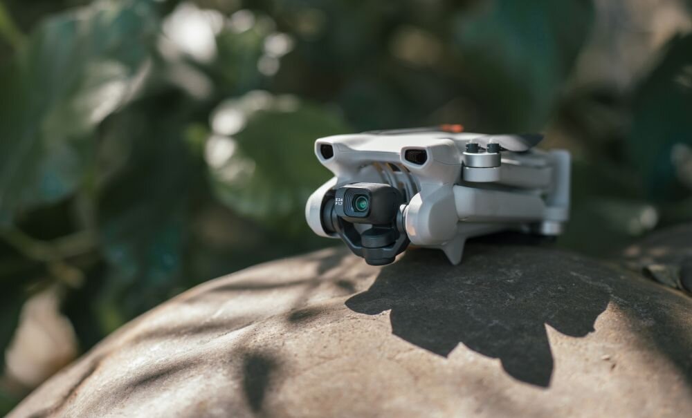 Na obrazie widoczny jest mały, składany dron z kamerą, umieszczony na kamieniu. W tle znajdują się rozmyte liście roślin.