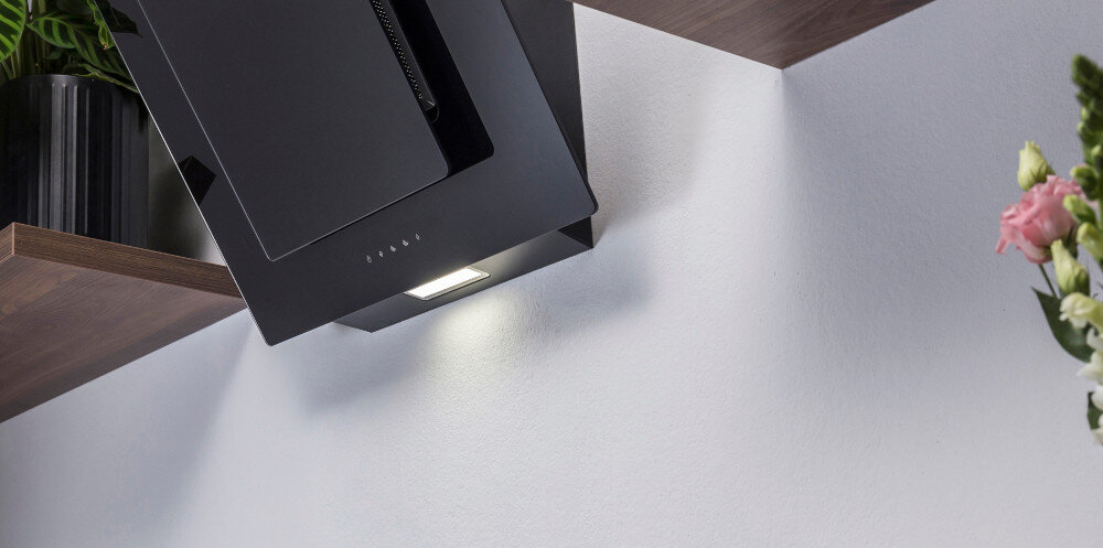 Czarny okap kuchenny zamontowany pod szafką, z włączonym oświetleniem. W tle widoczne są rośliny i kwiaty.