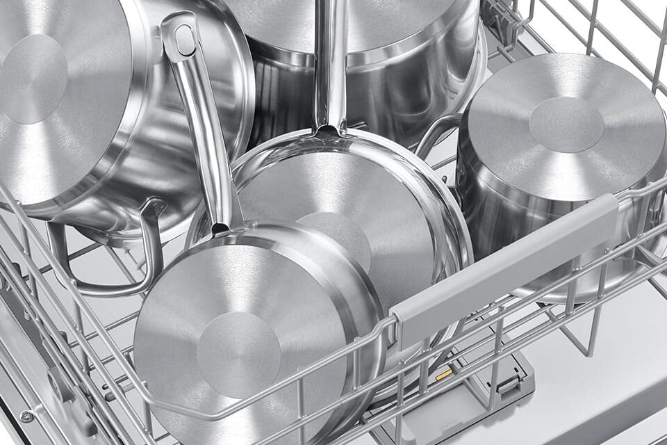 Wnętrze zmywarki z widocznymi metalowymi garnkami ułożonymi w koszu. Garnki mają błyszczącą powierzchnię i różne rozmiary.