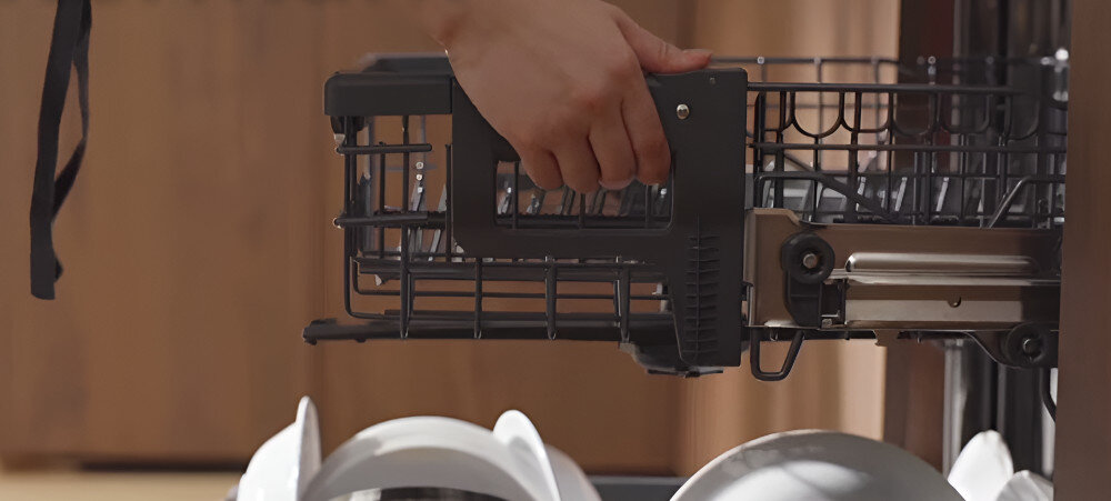 Osoba trzyma kosz na sztućce w zmywarce, w której widoczne są białe talerze. W tle znajduje się drewniana powierzchnia.