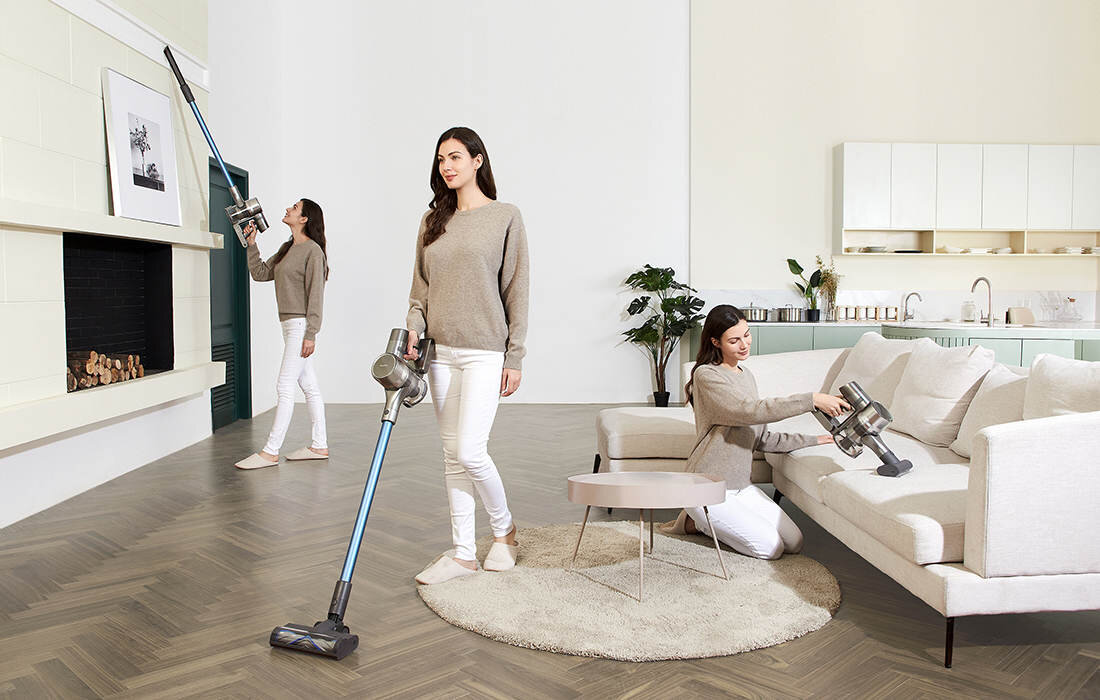 Trzy kobiety używają odkurzacza bezprzewodowego w nowoczesnym salonie z jasnymi meblami i drewnianą podłogą. W tle widoczna jest kuchnia z białymi szafkami i rośliną doniczkową.