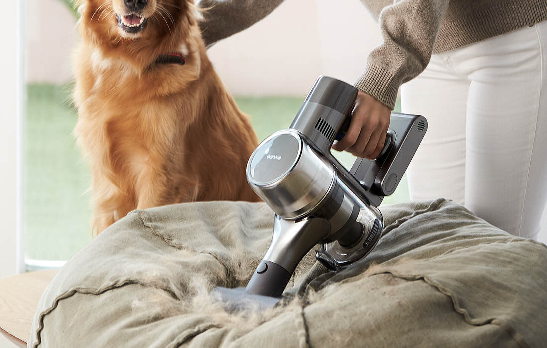 Osoba używa odkurzacza ręcznego do czyszczenia dużej poduszki z sierści. Obok znajduje się pies, a na odkurzaczu widoczne jest logo \'Dreame\'.
