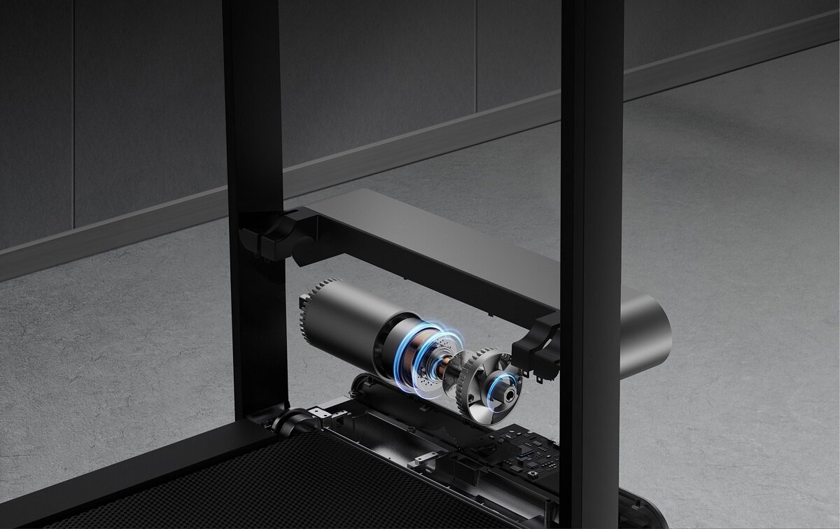 Obraz przedstawia mechanizm bieżni elektrycznej z widocznymi elementami konstrukcyjnymi i częściami wewnętrznymi. Tło jest jednolite, w szarym kolorze.