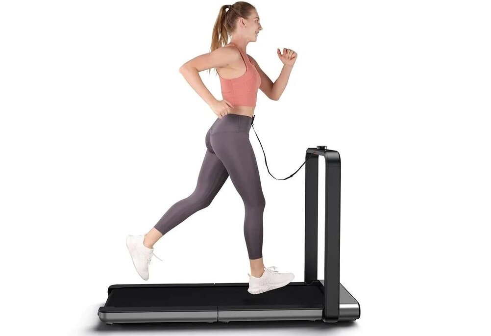 Osoba biegnie na bieżni elektrycznej z czarną podstawą i pionowym uchwytem. Bieżnia jest nowoczesna i kompaktowa.