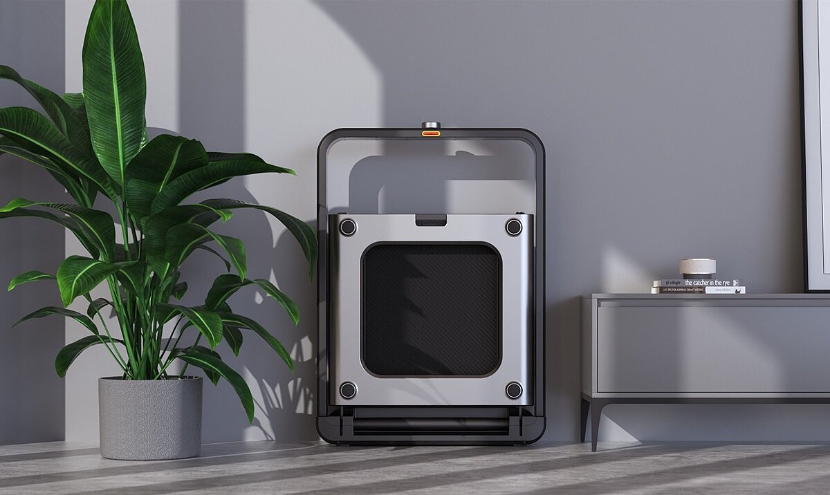 Na obrazie znajduje się złożona bieżnia elektryczna ustawiona pionowo obok rośliny doniczkowej i szafki. W tle widać szarą ścianę i podłogę z jasnymi panelami.