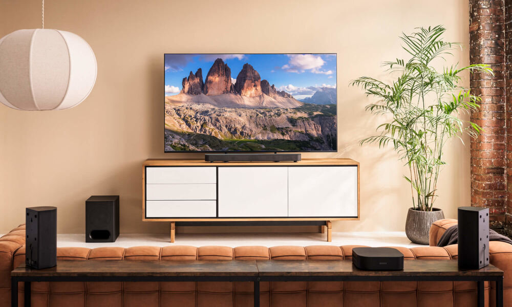 W salonie znajduje się telewizor na białej szafce, z widokiem na górski krajobraz. Przed szafką ustawiony jest soundbar, a po bokach widoczne są głośniki i subwoofer.