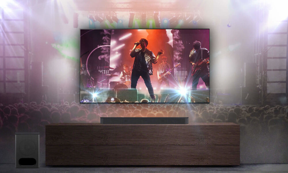 Na obrazie widać telewizor wyświetlający koncert, ustawiony na drewnianej szafce. Przed telewizorem znajduje się soundbar, a obok szafki widoczny jest głośnik niskotonowy.