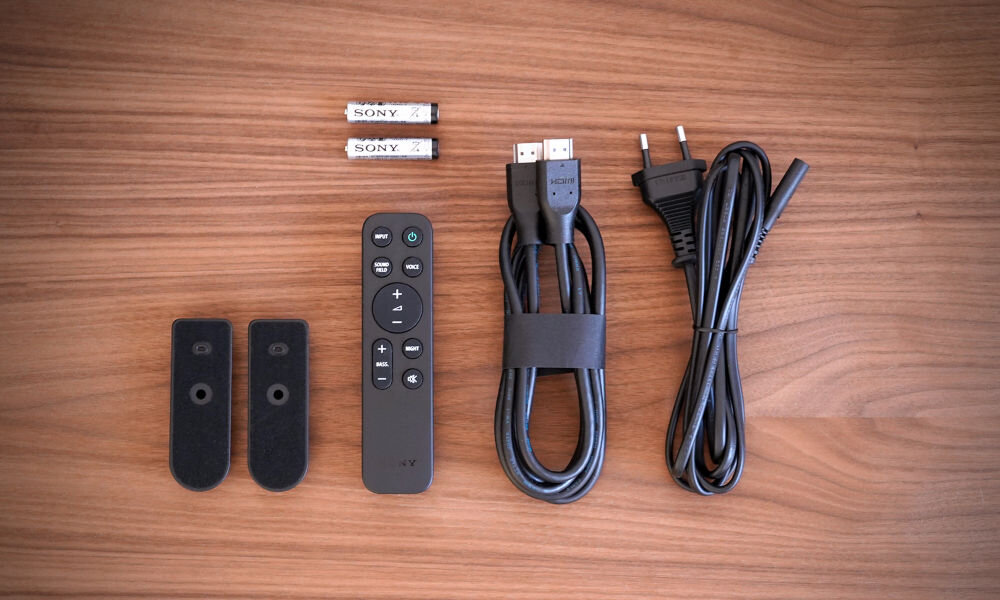 Na drewnianym tle leżą dwa piloty, dwa kable HDMI, kabel zasilający, dwa uchwyty montażowe oraz dwie baterie z logo SONY.