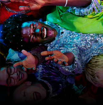 Grupa osób leży na podłodze, uśmiechając się i wyciągając ręce do góry. Osoby są ubrane w kolorowe, błyszczące stroje.