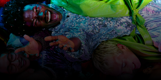 Na obrazie widać grupę osób w kolorowych ubraniach, leżących blisko siebie i uśmiechających się. Tło jest ciemne, co podkreśla jasne kolory ubrań.