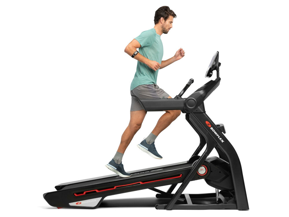 Osoba biegnie na czarnej bieżni elektrycznej z logo \'BOWFLEX\' na ramie. Bieżnia jest ustawiona pod kątem, a na panelu znajduje się ekran.