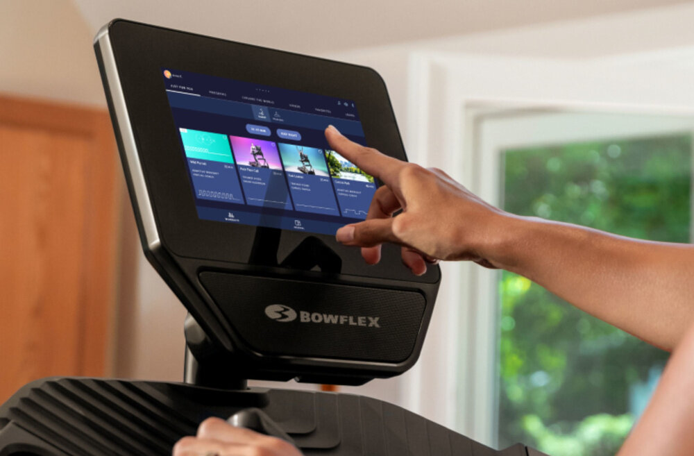 Osoba dotyka ekranu dotykowego na bieżni z widocznym logo \'BOWFLEX\'. Na ekranie wyświetlane są różne programy treningowe.