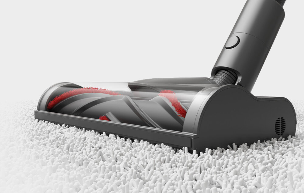 Na obrazie widoczna jest głowica odkurzacza z przezroczystą obudową, przez którą widać czerwone i szare elementy szczotki. Głowica znajduje się na białym, puszystym dywanie.
