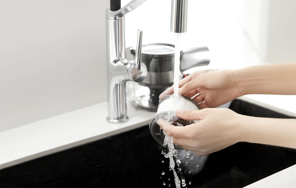 Osoba myje filtr pod bieżącą wodą w zlewie kuchennym. W tle widoczny jest metalowy kran i część urządzenia kuchennego.