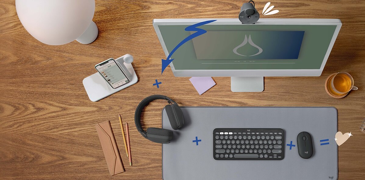 Na drewnianym biurku znajduje się monitor, klawiatura, mysz, słuchawki, telefon na podstawce, lampka, filiżanka z napojem oraz kilka kopert i ołówków. Klawiatura ma ciemny kolor i okrągłe klawisze, a obok niej leży mysz o podobnym designie.