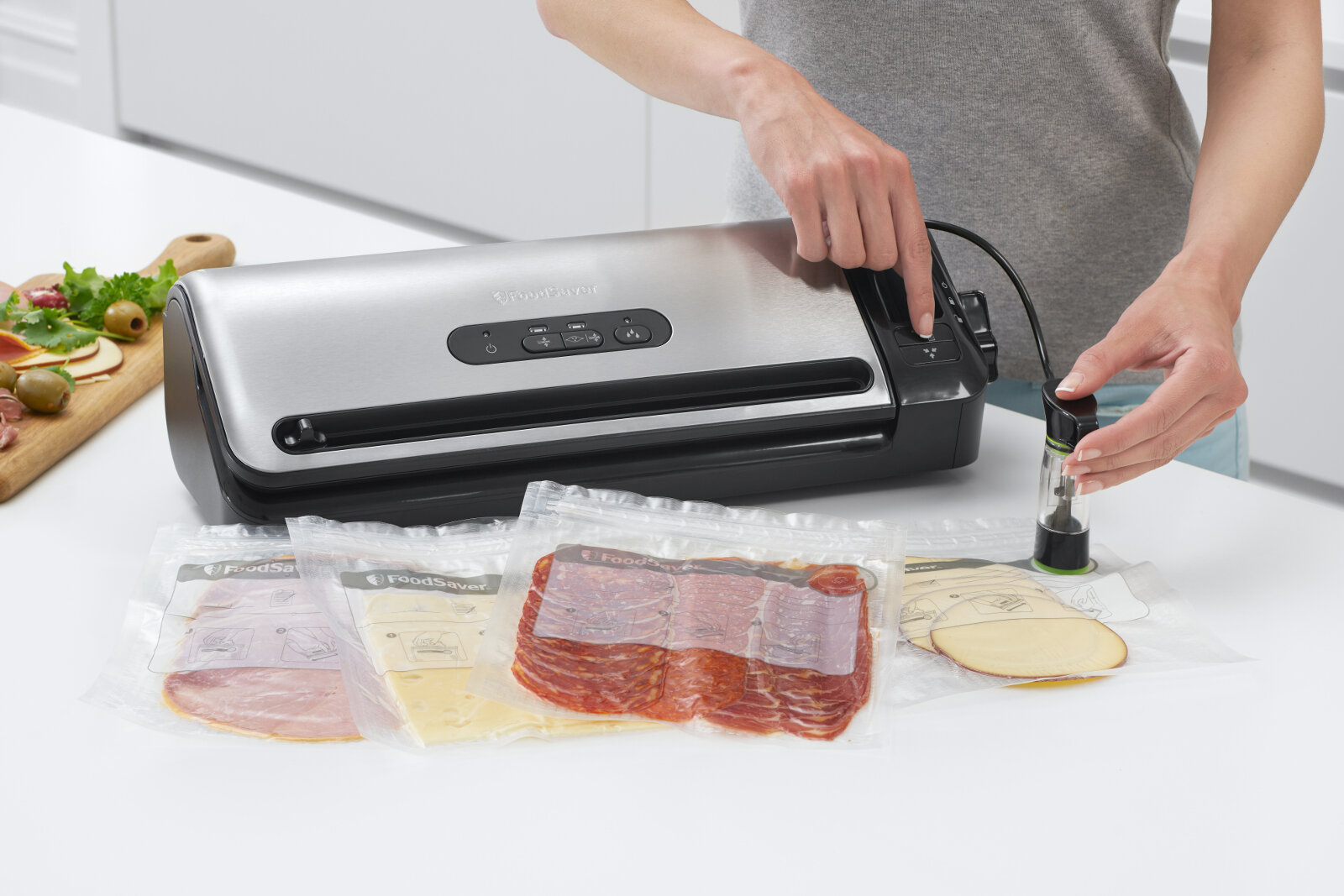 Osoba obsługuje zgrzewarkę próżniową, na blacie leżą zapakowane próżniowo produkty spożywcze. Na urządzeniu widoczne jest logo \'FoodSaver\'.