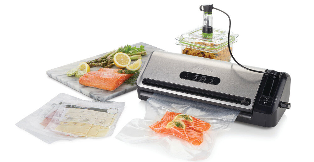 Na obrazie znajduje się zgrzewarka próżniowa z logo \'FoodSaver\', obok której leżą zapakowane próżniowo produkty spożywcze, takie jak łosoś i sery. W tle widoczny jest pojemnik z jedzeniem oraz deska z łososiem i szparagami.