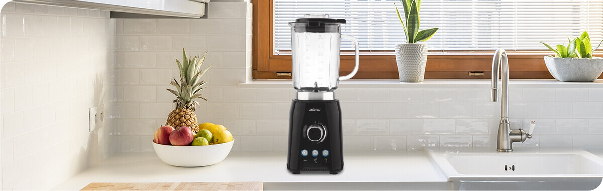 Na blacie kuchennym stoi czarny blender kielichowy marki ZELMER z pokrętłem i trzema przyciskami. Obok znajduje się miska z owocami oraz zlew, a w tle widać okno z roletą.