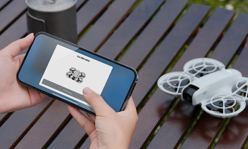 Osoba trzyma smartfon z wyświetlonym obrazem drona i tekstem \'DJI NEO 6054\'. Obok na stole znajduje się biały dron z osłonami na śmigła.