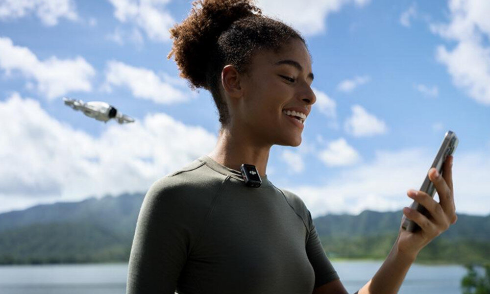 Osoba trzymająca smartfon, w tle widoczny jest dron na tle górskiego krajobrazu i nieba z chmurami.