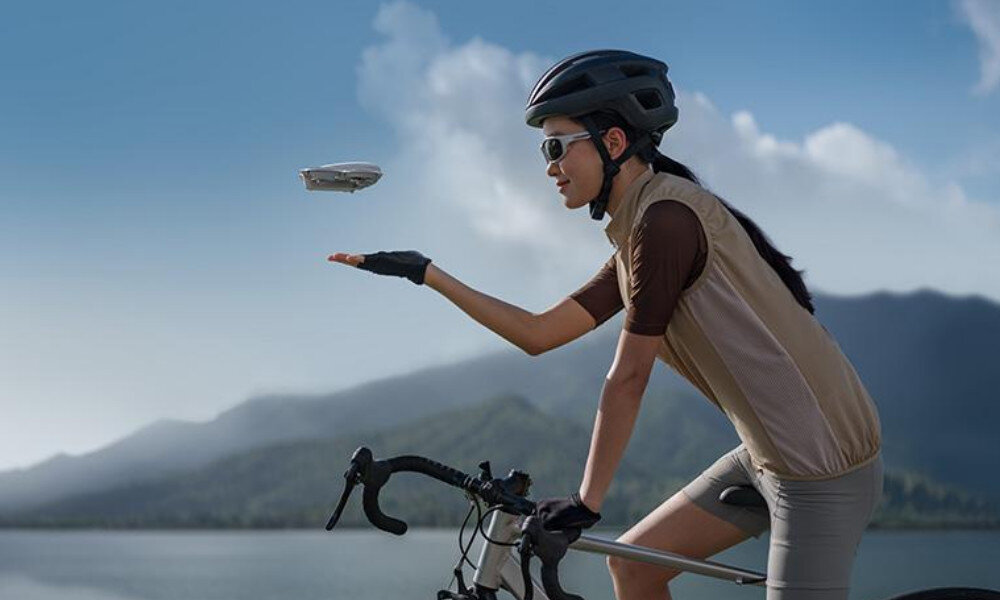 Osoba na rowerze trzyma rękę w górze, a nad nią unosi się mały dron. W tle widać góry i niebo z chmurami.