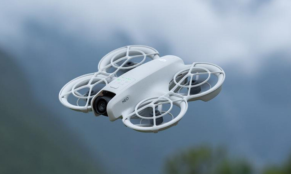 Na obrazie widoczny jest biały dron z czterema osłonami na śmigła, unoszący się w powietrzu na tle górskiego krajobrazu. Na dronie znajduje się napis \'NEO\'.