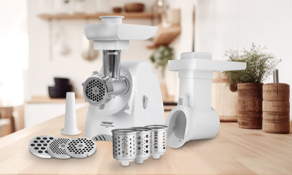 Na drewnianym blacie kuchennym znajduje się maszynka do mięsa z kilkoma wymiennymi tarczami i nasadkami. Widoczne jest logo \'Zelmer\' na urządzeniu.