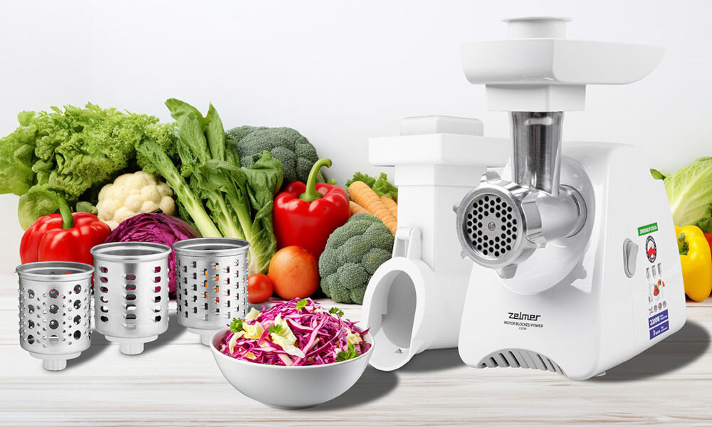 Na obrazie znajduje się maszynka do mięsa z logo \'zelmer\', obok której leżą różne warzywa oraz trzy metalowe tarki. Przed maszynką stoi miska z poszatkowaną kapustą.