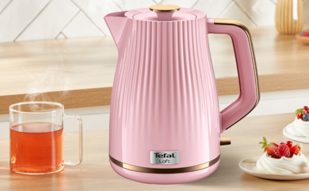 Różowy czajnik z logo \'Tefal Loft\' stoi na blacie kuchennym obok szklanki z herbatą i talerzyka z deserem.