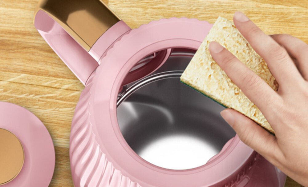 Różowy czajnik z otwartą pokrywą, wewnątrz którego widać metaliczne wnętrze. Obok czajnika znajduje się ręka trzymająca gąbkę, a całość leży na drewnianej powierzchni.