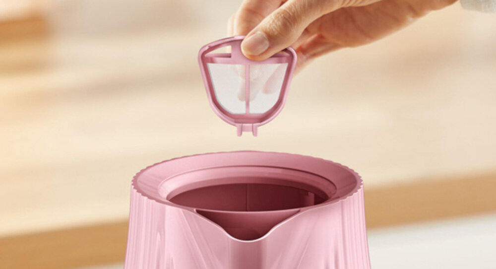 Różowy czajnik z otwartą pokrywą, nad którym unosi się ręka trzymająca filtr. Czajnik ma żebrowaną powierzchnię i szeroki otwór wlewowy.