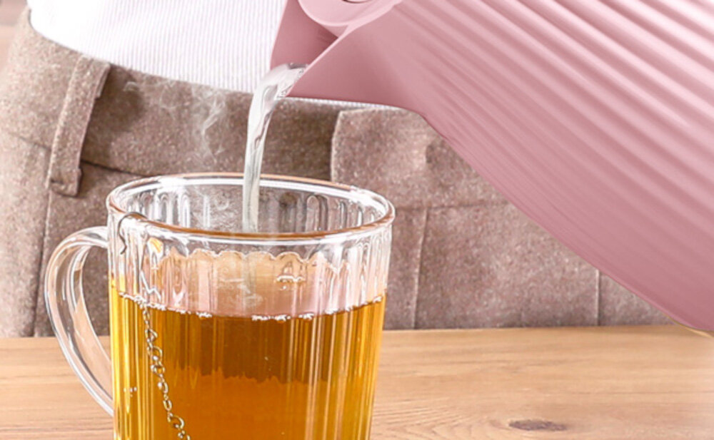 Różowy czajnik nalewa gorącą wodę do przezroczystej szklanki z herbatą na drewnianym stole.