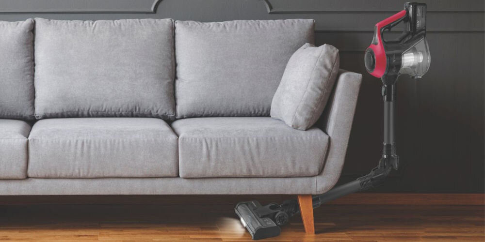 Odkurzacz w kolorze czarnym i czerwonym znajduje się pod szarą sofą na drewnianej podłodze. Urządzenie jest ustawione w pozycji pionowej z widocznym pojemnikiem na kurz.