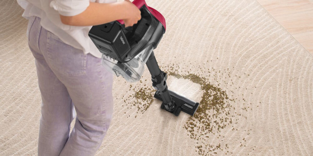 Osoba używa odkurzacza pionowego na jasnym dywanie, zbierając rozsypane drobinki. Na odkurzaczu widoczne jest logo \'Zelmer\'.