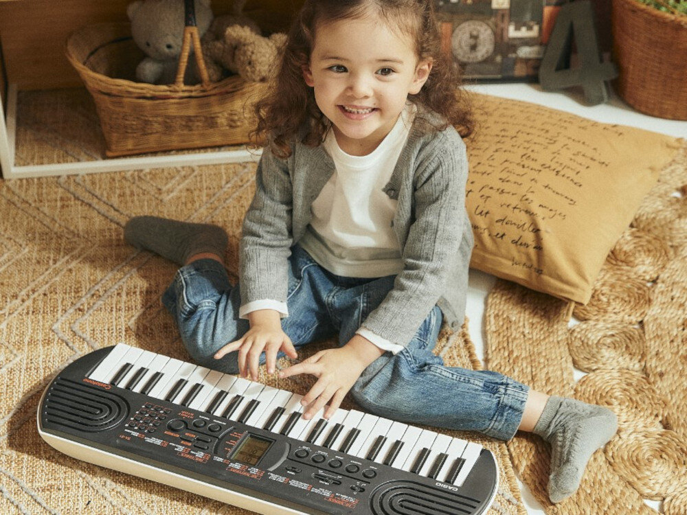 Dziecko siedzi na dywanie i gra na czarnym keyboardzie CASIO MU SA-81. W tle widoczne są kosz z pluszakami i poduszka z tekstem.