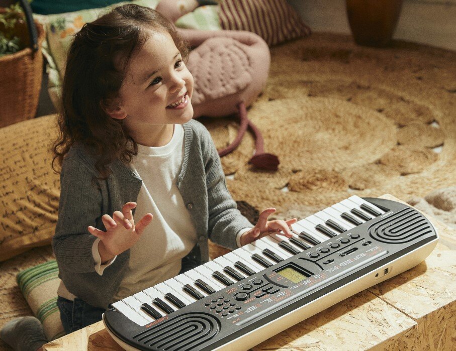 Dziecko siedzi przy keyboardzie CASIO MU SA-81 Czarny, który leży na drewnianej powierzchni. W tle widoczne są poduszki i dywan.