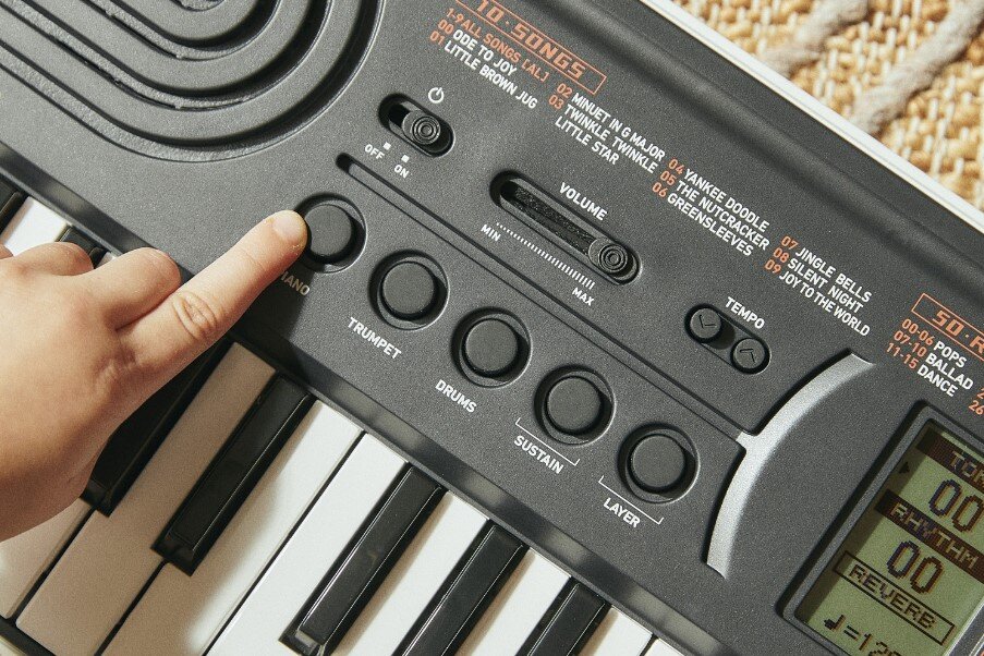 Na zdjęciu widać fragment czarnego keyboardu z widocznymi przyciskami i pokrętłami, a także wyświetlaczem LCD. Dłoń dziecka naciska jeden z przycisków oznaczony jako \'PIANO\'.